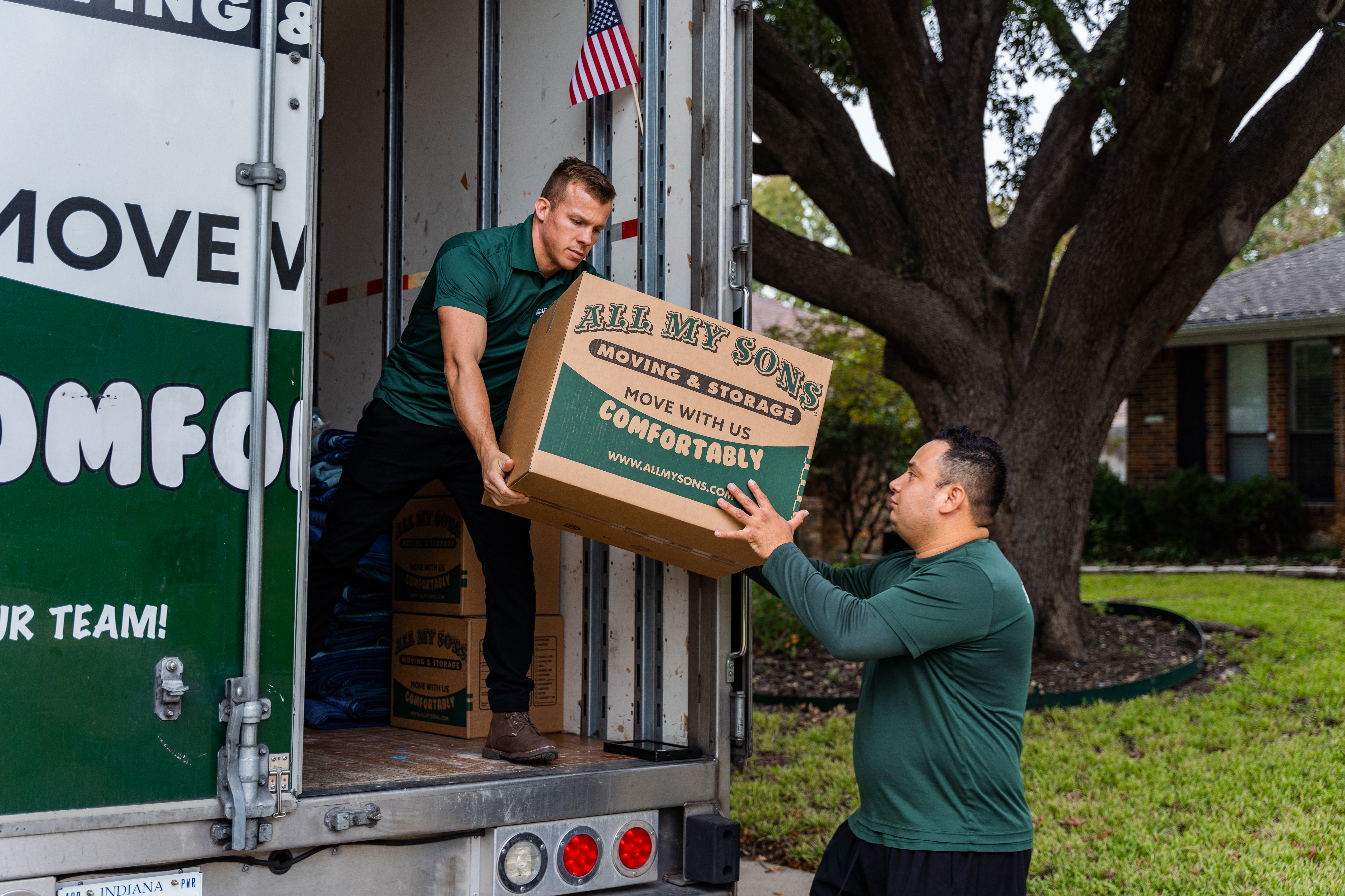 Two professional movers from All My Sons unload a moving box from the back of a moving truck.