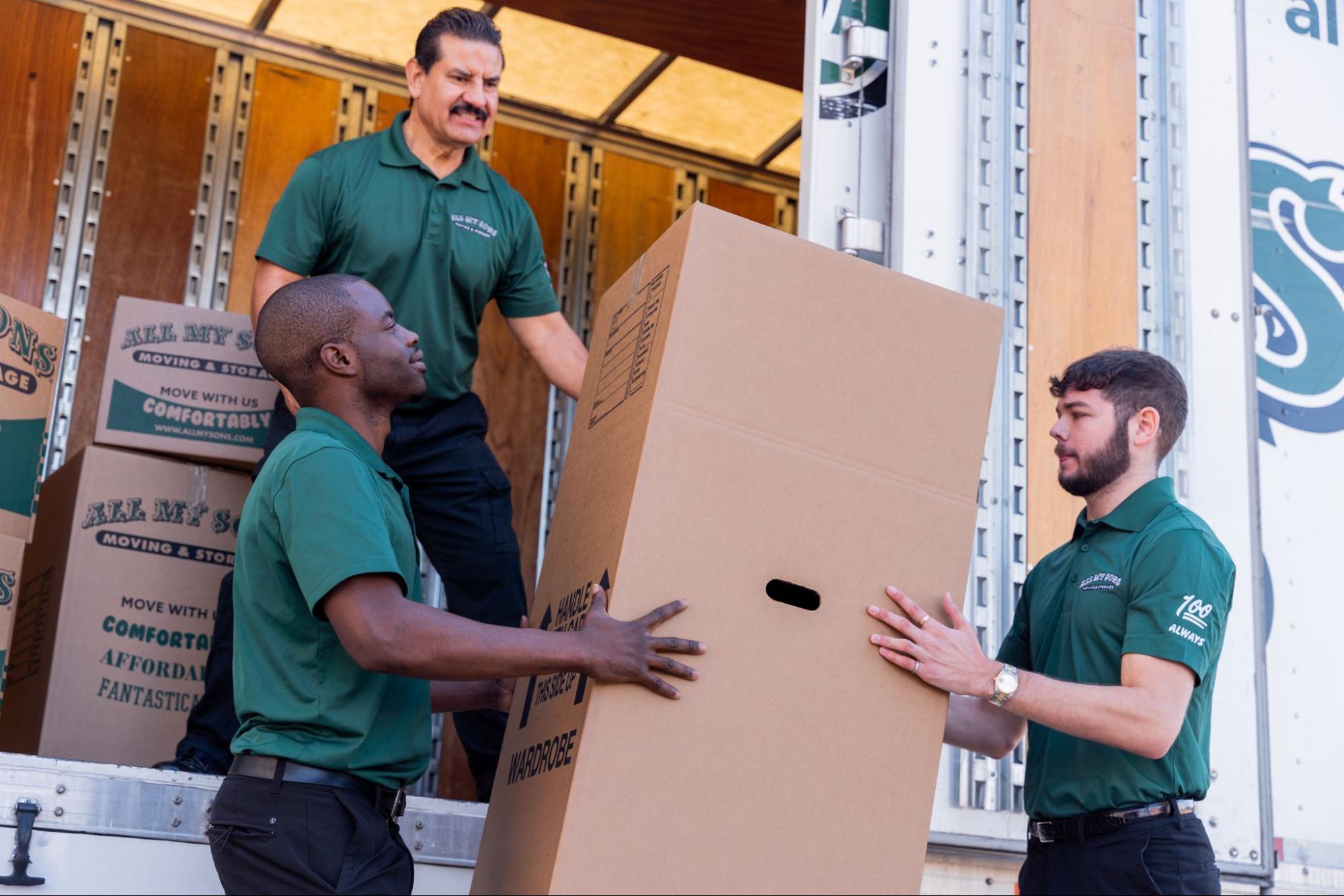 Three All My Sons professional movers lift a wardrobe box into the back of a truck.